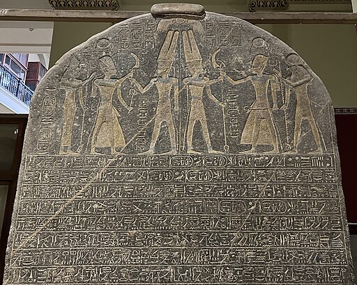 A close-up photograph of a tall, rounded dark granite stele (ancient Egyptian stone slab) covered in densely packed hieroglyphic